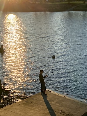 A person stands on a concrete platform with a fishing rod in hand, casting a line into a sunlit body of water. The sun reflects off the water, creating a golden path across the surface. Another figure is partially visible in the water, possibly wading.