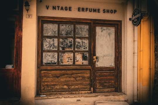 A rustic storefront with a weathered wooden door and window frames. The shop sign reads 'Vintage Perfume Shop' with some letters missing. The windows appear old and somewhat dirty, with an aged appearance. The surrounding wall is cream-colored, and there's a small black sign with ornate details hanging to the side.
