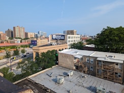 A cityscape featuring a mix of residential and commercial buildings. In the foreground, there are flat-roofed apartment buildings with wooden staircases and a view of the street below. Midway into the image, an older, ornate building with arches is visible. To the left, the street is lined with cars and bordered by trees and road signs. A large billboard advertises legal services. In the background, high-rise buildings dominate the skyline under a clear blue sky.