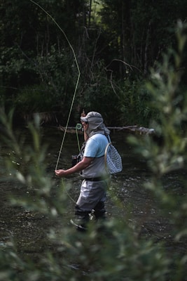 A person stands in a shallow stream, engaged in fly fishing. They are wearing waders, a light blue shirt, and a hat with a neck cover. A fishing net is attached to their back and they have a fly fishing rod in hand, with the line cast in the air. Surrounding the stream, dense green foliage creates a tranquil and secluded atmosphere.