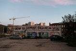A graffiti-covered wall offers tips on supporting Ukraine on social media, including messages like double-checking sources and posting important information. Two parked cars are in the foreground, while cranes and buildings fill the background.