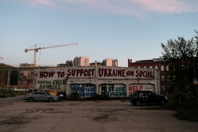 A graffiti-covered wall offers tips on supporting Ukraine on social media, including messages like double-checking sources and posting important information. Two parked cars are in the foreground, while cranes and buildings fill the background.