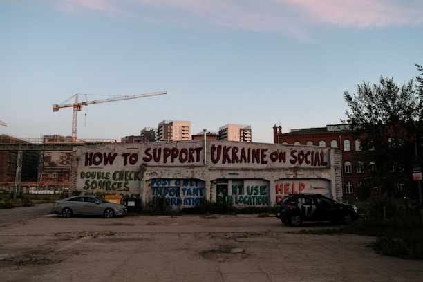 A graffiti-covered wall offers tips on supporting Ukraine on social media, including messages like double-checking sources and posting important information. Two parked cars are in the foreground, while cranes and buildings fill the background.