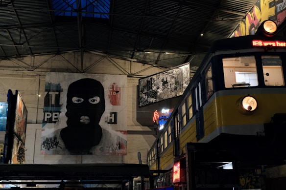 An indoor setting with urban, grunge-style artwork, including a large mural of a person wearing a balaclava. To the right, there is a train car with a neon sign inside. The space appears to be industrial, with exposed roofing and various other artworks and graffiti visible.