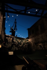 An urban skate park scene at dusk, enclosed by old brick buildings and modern graffiti art. Strings of lights create a warm atmosphere in the otherwise dimly-lit setting, and a person is performing a trick on a skateboard. The sky is a deep blue, contrasting with the silhouettes of the structures.