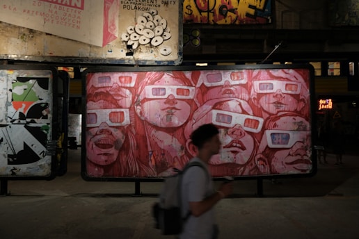 A street art installation featuring a large mural of faces wearing 3D glasses, drawn in shades of red. Above the mural, multiple old posters with various designs and text are visible. The scene is dimly lit, suggesting an urban, gritty environment. A person is walking in front of the mural, slightly blurred, adding a sense of motion.