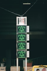 A vertical electronic sign with green illuminated characters, displaying availability information in Japanese. It is mounted on a pole against a dark background, with several small potted plants at the base. The scene includes additional signage with text in Japanese.