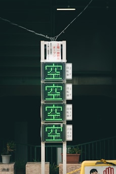 A vertical electronic sign with green illuminated characters, displaying availability information in Japanese. It is mounted on a pole against a dark background, with several small potted plants at the base. The scene includes additional signage with text in Japanese.