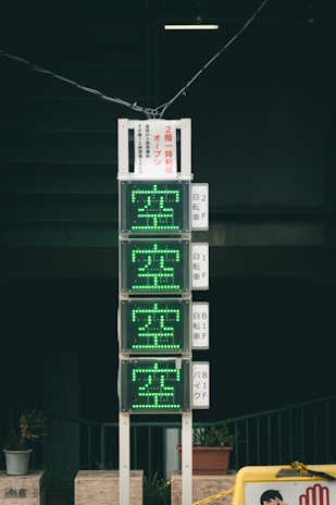 A vertical electronic sign with green illuminated characters, displaying availability information in Japanese. It is mounted on a pole against a dark background, with several small potted plants at the base. The scene includes additional signage with text in Japanese.