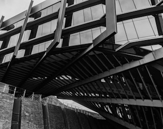 A modern architectural structure featuring angular lines and large glass windows. Multiple metal beams and geometric shapes create a complex pattern, with reflections visible in the glass. The structure seems to rise above a brick wall, adding an industrial feel.
