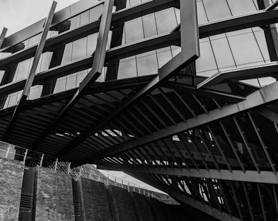 A modern architectural structure featuring angular lines and large glass windows. Multiple metal beams and geometric shapes create a complex pattern, with reflections visible in the glass. The structure seems to rise above a brick wall, adding an industrial feel.