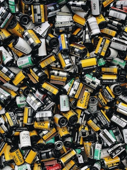 A large assortment of film canisters piled together, featuring various brands and ISO numbers. The canisters are cylindrical, with a mix of vibrant colors such as yellow, green, and black, each with distinctive labeling.