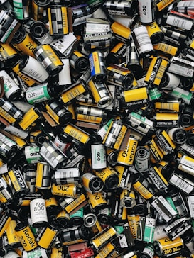 A large assortment of film canisters piled together, featuring various brands and ISO numbers. The canisters are cylindrical, with a mix of vibrant colors such as yellow, green, and black, each with distinctive labeling.