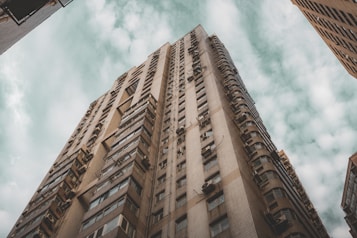A tall, residential skyscraper viewed from below against a sky filled with scattered clouds. The building has a grid-like facade with numerous windows and air conditioning units attached, creating a sense of urban density.