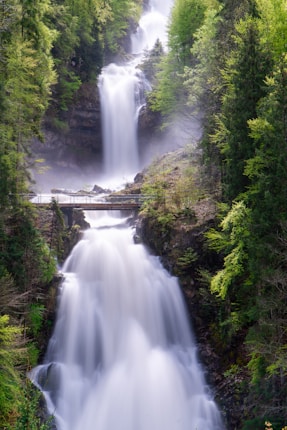 A majestic multi-tiered waterfall cascades down a rocky cliff, surrounded by lush green trees. Mist rises from the water as it falls, creating a serene and refreshing atmosphere. A bridge crosses over the waterfall, blending harmoniously into the natural surroundings.