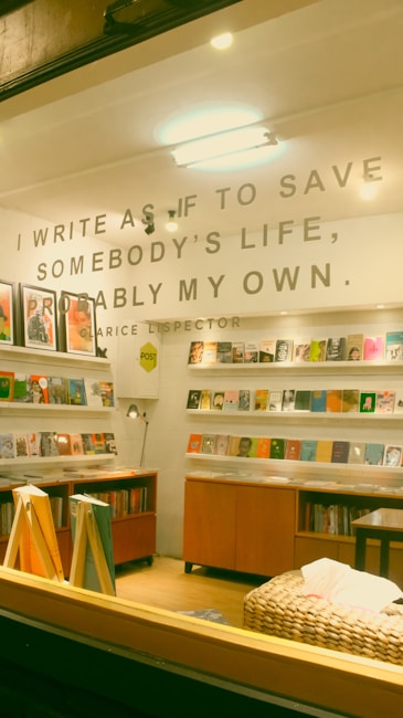 A cozy indoor space featuring a well-organized bookshelf filled with various books. Above the bookshelves, there's a quote written on the wall: 'I write as if to save somebody&rsquo;s life, probably my own. - Clarice Lispector'. The room is warmly lit with a mix of wooden furniture and a cushioned area in the foreground.