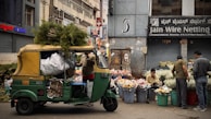A bustling street market scene featuring an auto rickshaw loaded with greenery and sacks, parked in front of a flower stall. Vendors sit and stand beside large buckets filled with assorted flowers. The background displays urban storefronts with signage.