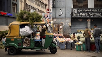 A bustling street market scene featuring an auto rickshaw loaded with greenery and sacks, parked in front of a flower stall. Vendors sit and stand beside large buckets filled with assorted flowers. The background displays urban storefronts with signage.