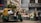 A bustling street market scene featuring an auto rickshaw loaded with greenery and sacks, parked in front of a flower stall. Vendors sit and stand beside large buckets filled with assorted flowers. The background displays urban storefronts with signage.