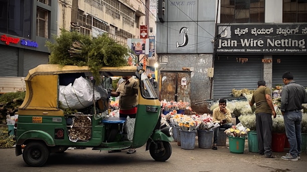 A bustling street market scene featuring an auto rickshaw loaded with greenery and sacks, parked in front of a flower stall. Vendors sit and stand beside large buckets filled with assorted flowers. The background displays urban storefronts with signage.