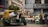 A bustling street market scene featuring an auto rickshaw loaded with greenery and sacks, parked in front of a flower stall. Vendors sit and stand beside large buckets filled with assorted flowers. The background displays urban storefronts with signage.