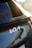 A close-up view of the rear section of a car displaying the logo of the automotive brand Kia. The car has a glossy, dark-colored surface that reflects the surroundings, including parts of a structure with an abstract pattern.