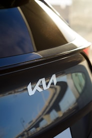 A close-up view of the rear section of a car displaying the logo of the automotive brand Kia. The car has a glossy, dark-colored surface that reflects the surroundings, including parts of a structure with an abstract pattern.