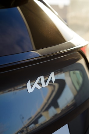 A close-up view of the rear section of a car displaying the logo of the automotive brand Kia. The car has a glossy, dark-colored surface that reflects the surroundings, including parts of a structure with an abstract pattern.