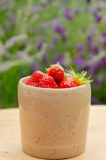 A speckled beige ceramic cup is filled with fresh red strawberries, set against a blurred background of vibrant green foliage and purple flowers.