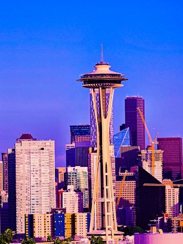 A vibrant cityscape featuring an iconic tower in the foreground, surrounded by modern high-rise buildings. Construction cranes are visible among the skyscrapers, indicating ongoing development. The image is saturated with vivid colors, enhancing the architectural features against a clear blue sky.