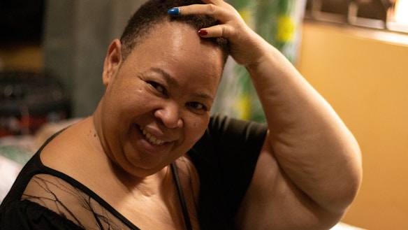 A person smiling warmly while touching their head with one hand. The person is wearing a dark top with sheer details and has painted nails in blue and red. The background features a blurred setting with soft lighting, including green and yellow elements.
