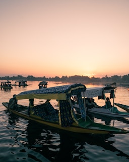 A serene lake scene at sunset with multiple traditional boats floating on the calm water. Each boat is adorned with colorful fabrics, and the silhouettes of people can be seen. The sky is a gradient of warm hues, and the distant silhouette of trees lines the horizon.