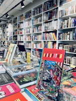 A bookstore interior filled with shelves of books arranged neatly. Several colorful books are displayed prominently on a table in the foreground, with one particular book featuring a vibrant and graphic cover standing upright.