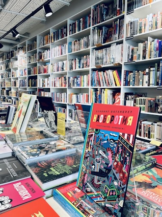 A bookstore interior filled with shelves of books arranged neatly. Several colorful books are displayed prominently on a table in the foreground, with one particular book featuring a vibrant and graphic cover standing upright.