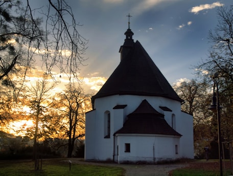 A small, white, round church with a dark, sloping roof set against a backdrop of a vibrant sunset. The sky is a mix of blue and warm golden tones, with bare tree branches framing the scene. A lamp post is visible on the right, and the grassy ground leads up to the church.
