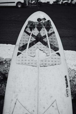 A monochrome photograph of a surfboard leaning against a rough stone surface. The surfboard has a traction pad with a triangular pattern near the tail fin area, and a leash cord is visible. The background shows a parked vehicle and a street, slightly out of focus.