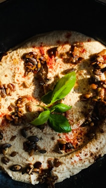 A close-up of a creamy dip garnished with green basil leaves and sprinkled with seeds and spices. The dish has a rich and textured appearance, with a scattering of red seasoning adding contrast to the light beige base.