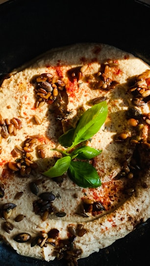 A close-up of a creamy dip garnished with green basil leaves and sprinkled with seeds and spices. The dish has a rich and textured appearance, with a scattering of red seasoning adding contrast to the light beige base.
