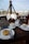 A cozy outdoor cafe setting featuring a wooden table with two cups of coffee and two desserts. The table setup includes plates, cutlery, and a small container for sugar packets. In the background, there's a glass panel with writing and a view of the surrounding outdoor area.