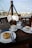 A cozy outdoor cafe setting featuring a wooden table with two cups of coffee and two desserts. The table setup includes plates, cutlery, and a small container for sugar packets. In the background, there's a glass panel with writing and a view of the surrounding outdoor area.