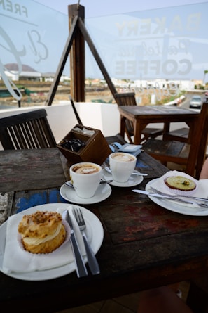 A cozy outdoor cafe setting featuring a wooden table with two cups of coffee and two desserts. The table setup includes plates, cutlery, and a small container for sugar packets. In the background, there's a glass panel with writing and a view of the surrounding outdoor area.