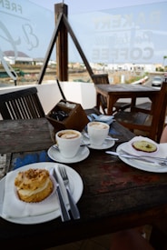 A cozy outdoor cafe setting featuring a wooden table with two cups of coffee and two desserts. The table setup includes plates, cutlery, and a small container for sugar packets. In the background, there's a glass panel with writing and a view of the surrounding outdoor area.
