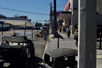 A street scene with a golf cart parked on the side of a busy road. There are a few people walking on the sidewalk near shops and a bicycle rental station with several bikes. The sky is clear and blue, and a sign reading 'VENICE' hangs over the street in the distance.