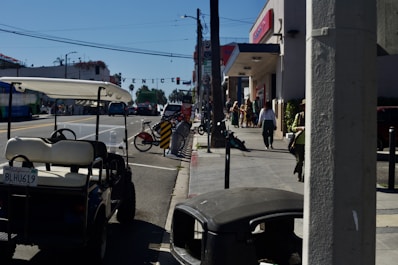 A street scene with a golf cart parked on the side of a busy road. There are a few people walking on the sidewalk near shops and a bicycle rental station with several bikes. The sky is clear and blue, and a sign reading 'VENICE' hangs over the street in the distance.