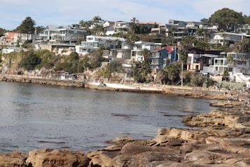 A coastal scene featuring a rocky shoreline and calm seawater. Modern houses are built on a hillside overlooking the ocean, surrounded by greenery and palm trees. The architecture of the homes is contemporary, with large glass windows. People are seen walking along a path near the water.