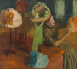 A woman dressed in period clothing is seated, examining a hat she holds in her hands. Around her are several other hats on stands, each with distinct designs and colors. The setting appears to be an interior space with a focus on fashion or millinery.