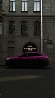 A vibrant purple SUV is parked on a street in front of a building with a gray stone facade. The building features large windows with an archway entrance displaying a neon sign with Cyrillic text. A street sign is also visible on the side of the building.