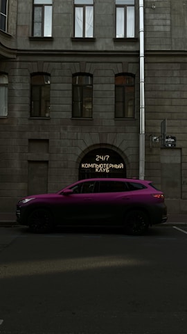 A vibrant purple SUV is parked on a street in front of a building with a gray stone facade. The building features large windows with an archway entrance displaying a neon sign with Cyrillic text. A street sign is also visible on the side of the building.