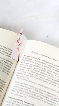 An open book on a light surface features a decorative bookmark with pink cherry blossoms. The text on the pages discusses Japanese cultural themes and philosophies, with visible sections about Shinto and the influence of Buddhism.