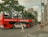 A red double-decker tour bus is driving down an urban street with a person walking across the road. The scene includes several buildings, a tall tree, and signs for tourist information and parking. The sky is partly cloudy.